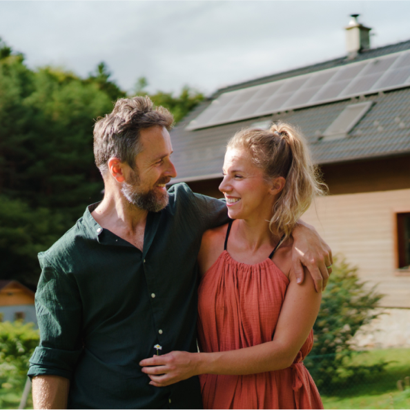 Man and woman standing in front of a house with solar panels. Man has his arm around the woman and they are both smiling.